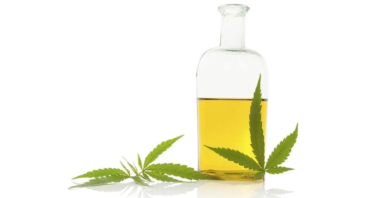 Bottle of cannabis oil with green cannabis leaves on a reflective white surface.
