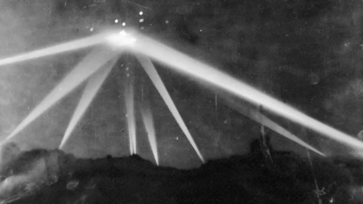 Historical black-and-white photograph of searchlights illuminating the night sky during a wartime event